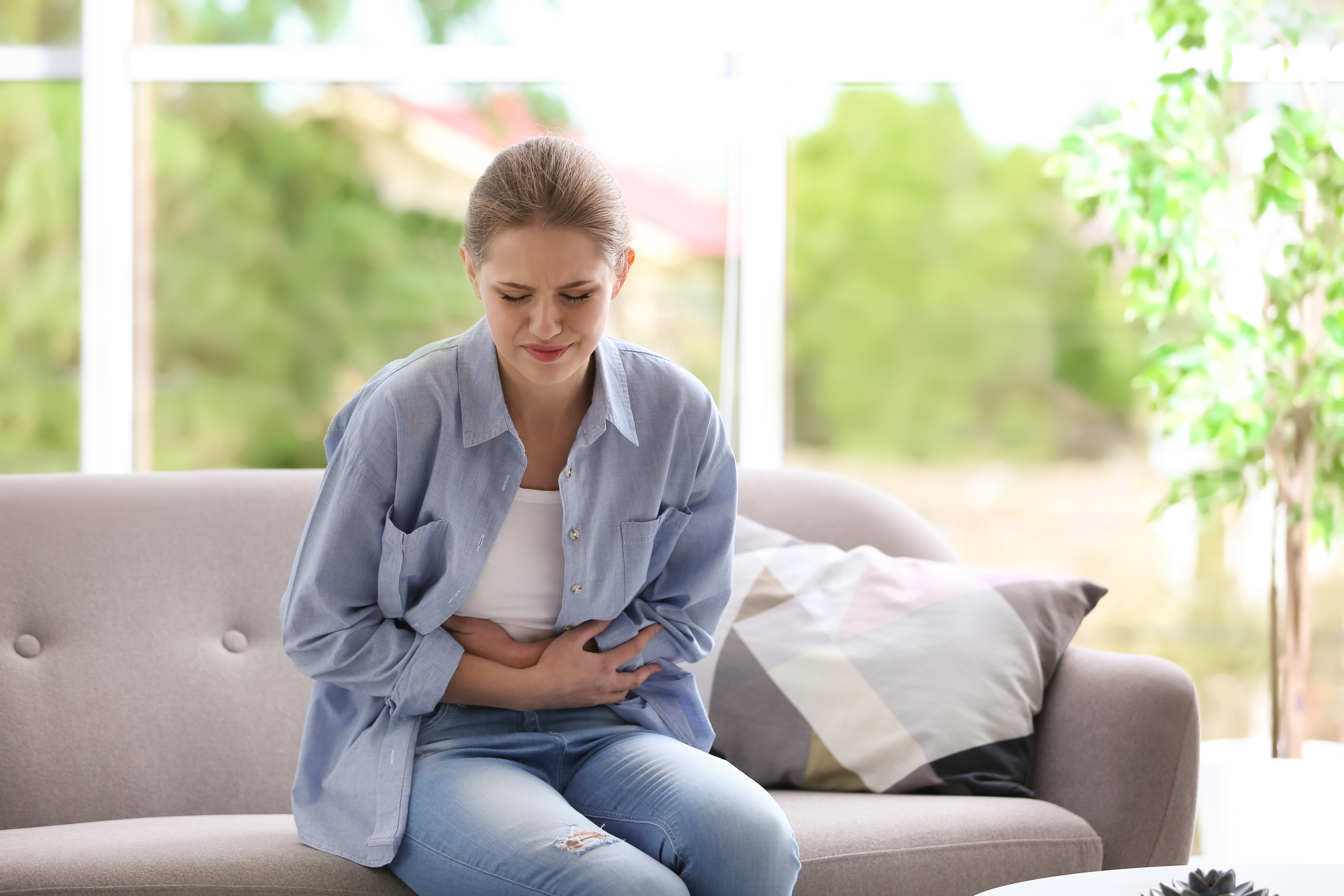 Mulher sentada em sofá e com mãos na barriga depois de sentir um dos sintomas da endometriose