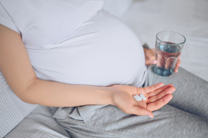 Mulher grávida segurando pílulas de remédios em uma das mãos com um copo de água em outra.
