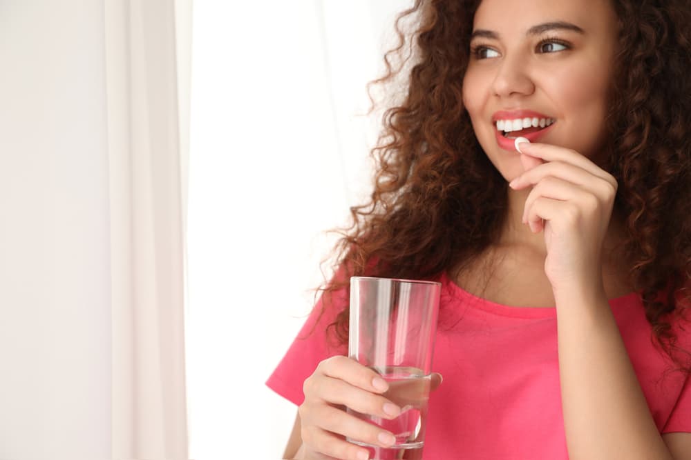 mulher com copo de água em uma mão e colocando comprimido na boca para representar reposição hormonal