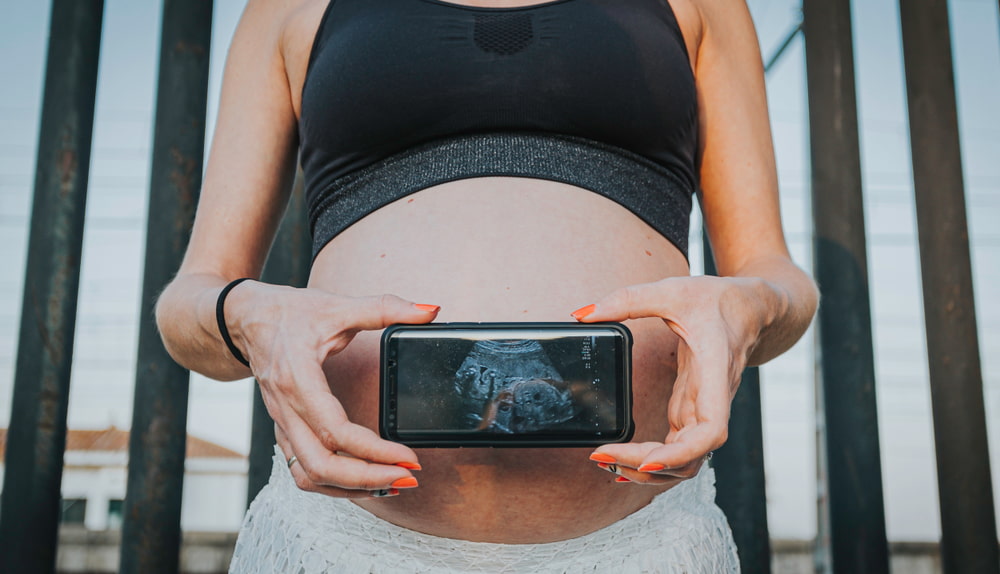Mulher grávida com um celular posicionado a frente da barriga com a imagem de um ultrassom.