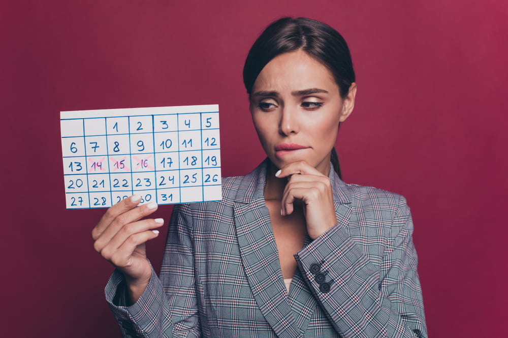 Mulher branca com um blazer cinza olhando para um calendário fértil com uma fisionomia de pensativa. O fundo da imagem é vinho.