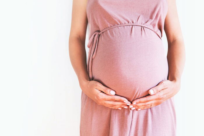 Mulher branca grávida com as mãos posicionadas abaixo da barriga.