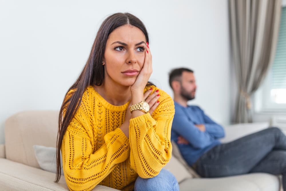 mulher sentada no sofá com cara de preocupação por conta de infertilidade sem causa aparente, marido está sentado ao fundo